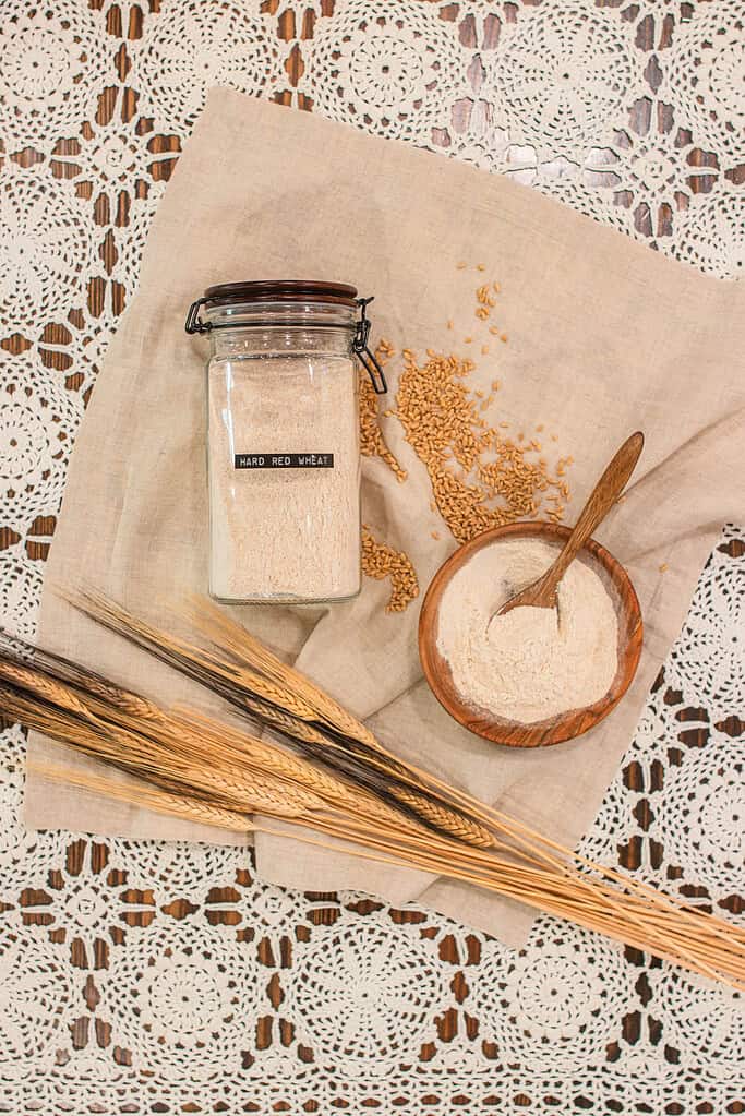 Glass jar labeled Hard Red Wheat filled with freshly milled flour, resting beside a wooden bowl of flour and spoon, loose wheat berries, and wheat stalks on linen over lace — demonstrating long-term flour storage in an airtight container.