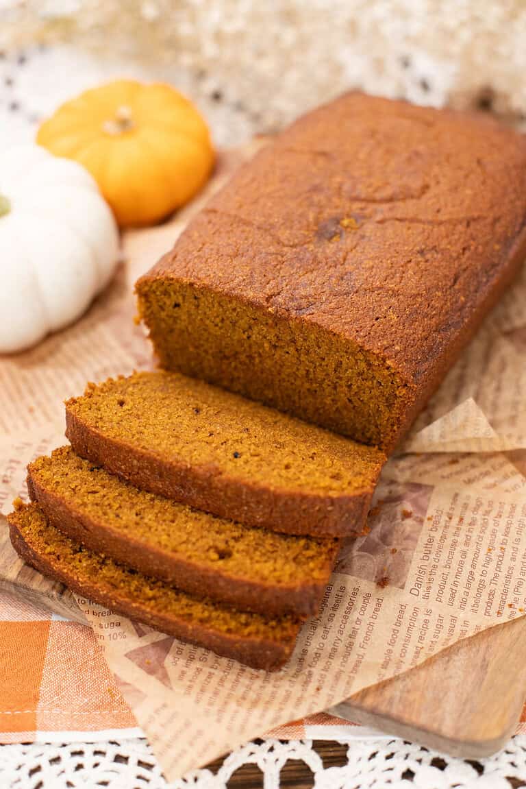 A freshly baked loaf of pumpkin bread, golden brown and moist, sliced to reveal its tender crumb. The loaf rests on parchment-style paper with mini pumpkins and fall décor in the background.
