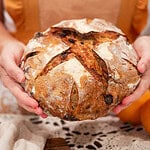 A woman in a warm-toned apron holding a rustic loaf of sourdough cranberry orange bread with a beautifully scored crust.