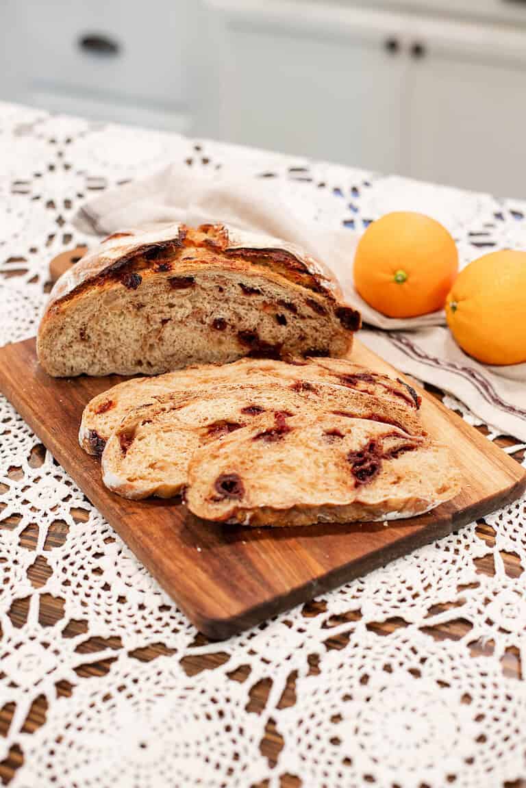 Sliced sourdough cranberry orange bread displayed on a wooden cutting board with oranges in the background, showing soft texture and cranberry swirls.