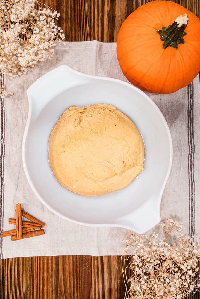 Pumpkin sourdough dough resting in a white mixing bowl beside a small pumpkin, cinnamon sticks, and dried flowers on a wooden table.