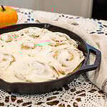 Freshly baked sourdough pumpkin cinnamon rolls in a cast-iron skillet topped with cream cheese frosting, set on a lace tablecloth with cinnamon sticks and small pumpkins for a cozy fall touch.