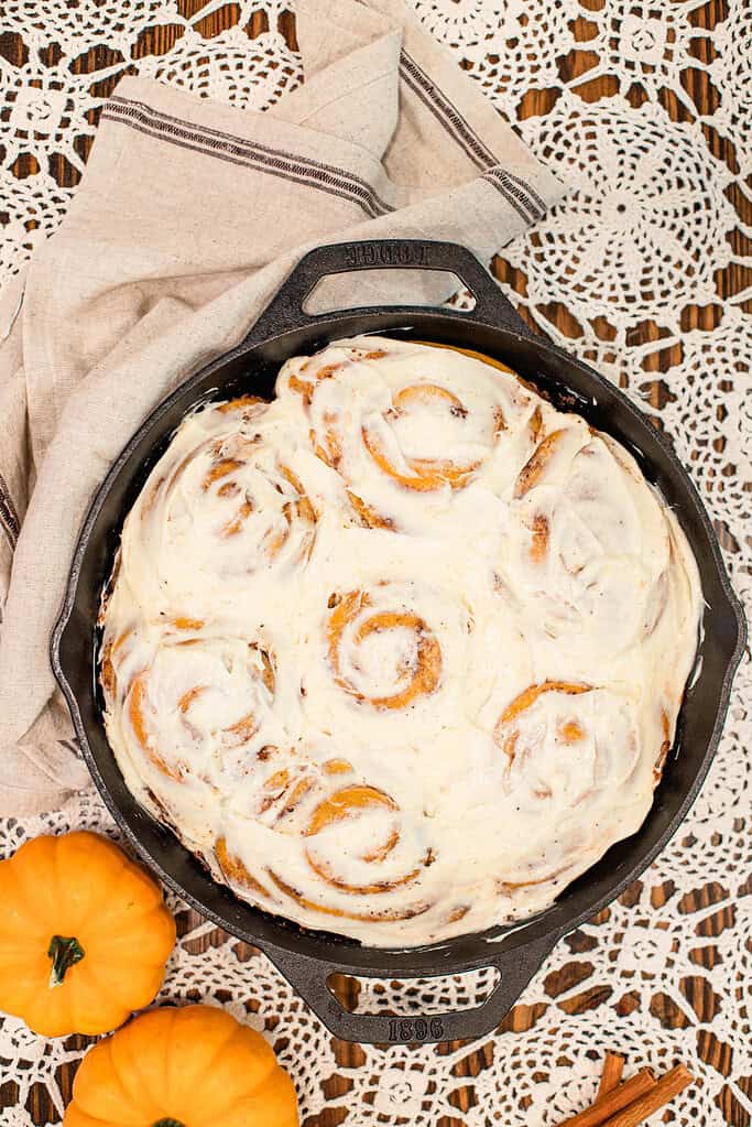 Freshly baked sourdough pumpkin cinnamon rolls in a cast-iron skillet, generously topped with creamy frosting, surrounded by pumpkins and fall decor.