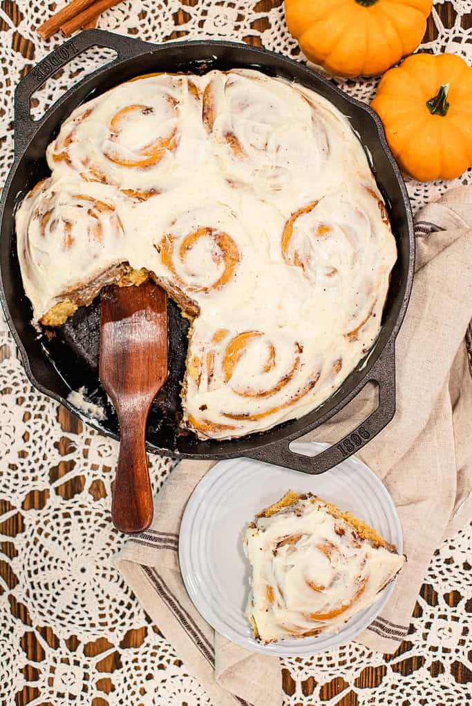 Cast-iron skillet filled with frosted sourdough pumpkin cinnamon rolls with one slice removed and served on a small white plate.