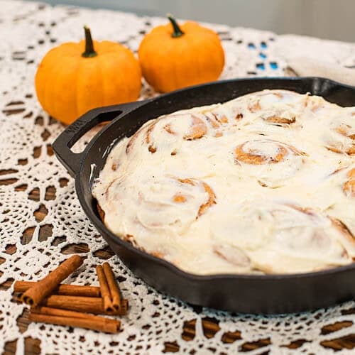Freshly baked sourdough pumpkin cinnamon rolls in a cast-iron skillet topped with cream cheese frosting, set on a lace tablecloth with cinnamon sticks and small pumpkins for a cozy fall touch.