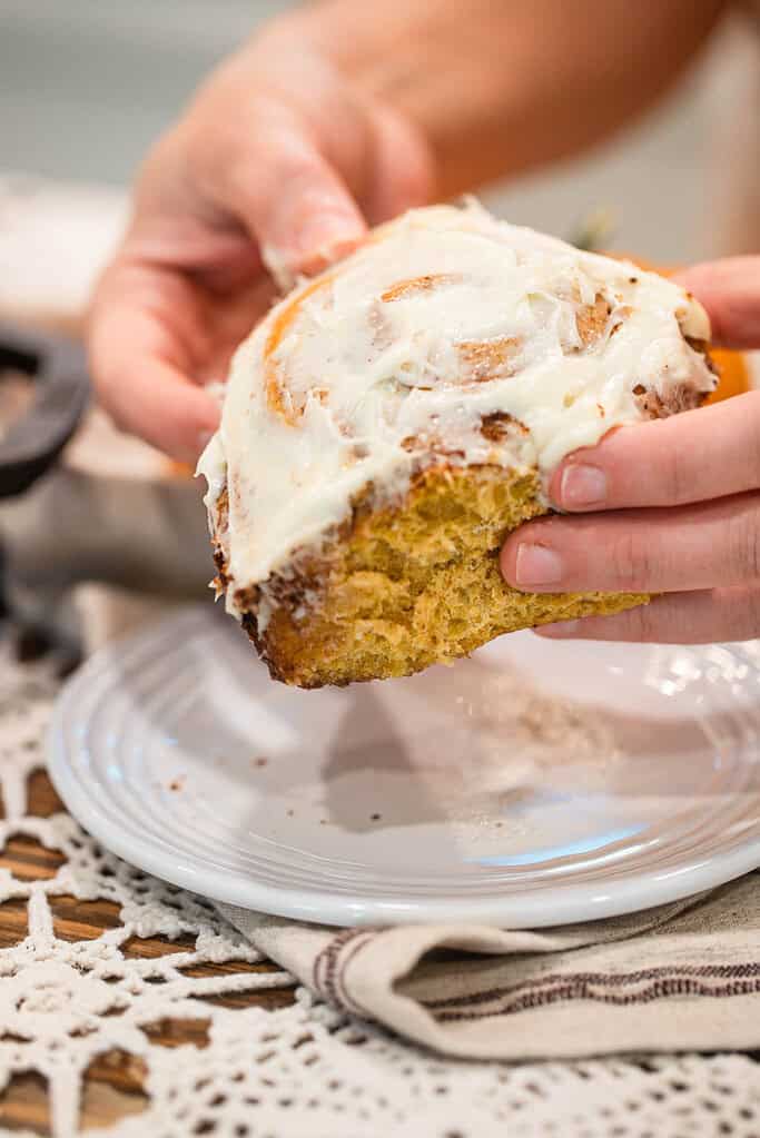 Hands holding a frosted sourdough pumpkin cinnamon roll above a white plate, showing the soft, fluffy pumpkin-spiced texture.