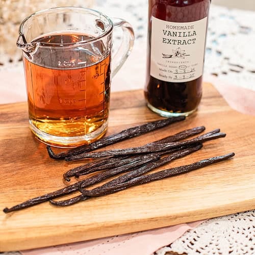 Vanilla beans on a wooden board next to a measuring cup of alcohol and a bottle of homemade vanilla extract.