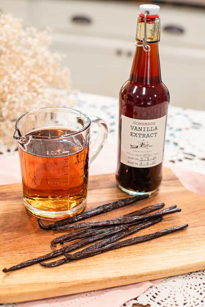 Vanilla beans on a wooden board next to a measuring cup of alcohol and a bottle of homemade vanilla extract.