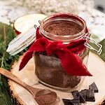 An open jar of homemade hot cocoa mix on a wooden slice with a wooden spoon and pieces of dark chocolate.