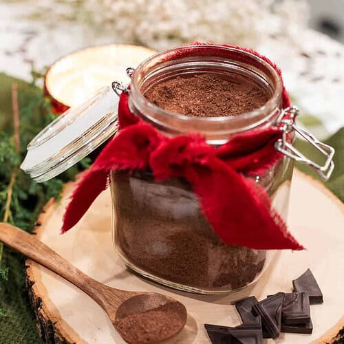 An open jar of homemade hot cocoa mix on a wooden slice with a wooden spoon and pieces of dark chocolate.