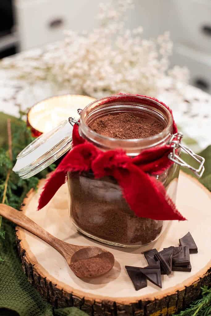 An open jar of homemade hot cocoa mix on a wooden slice with a wooden spoon and pieces of dark chocolate.