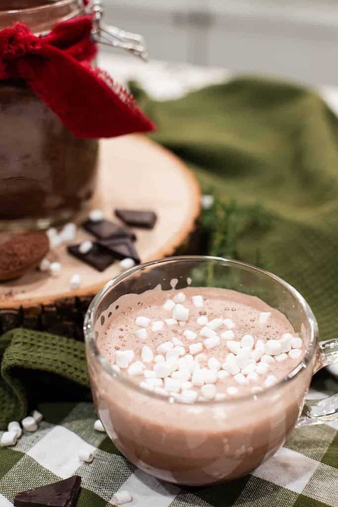 A clear glass mug of hot cocoa topped with mini marshmallows, sitting beside a jar of cocoa mix.