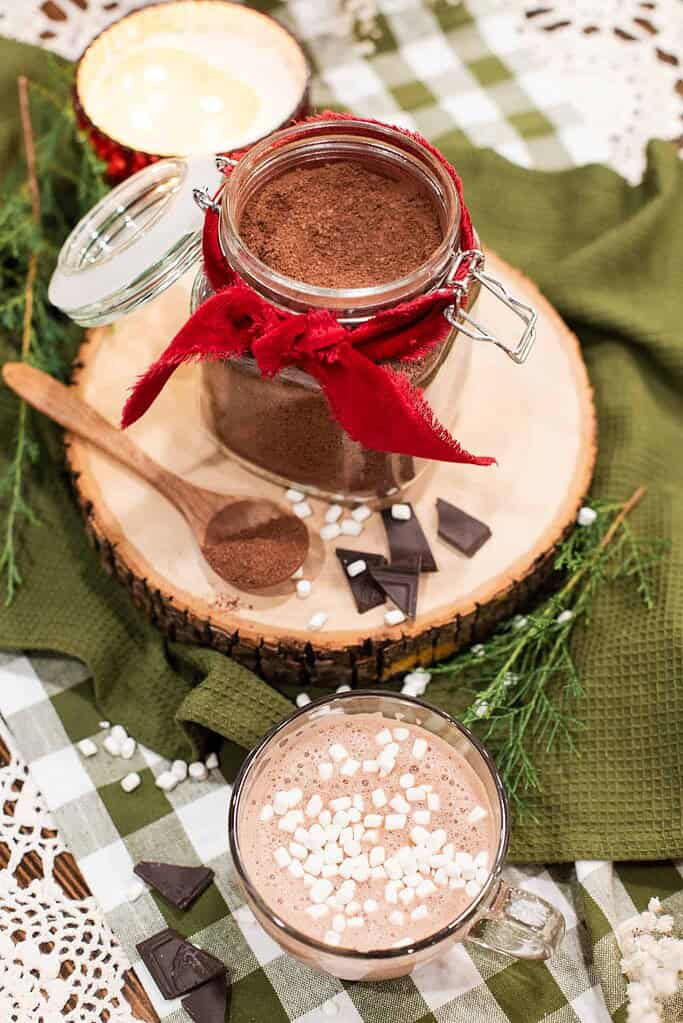 Overhead shot of a jar of hot cocoa mix with a spoon and chocolate pieces, beside a mug of hot cocoa topped with marshmallows.