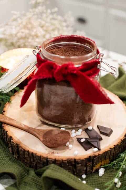 A jar of hot cocoa mix decorated with a red ribbon, shown with a wooden spoon and dark chocolate pieces.