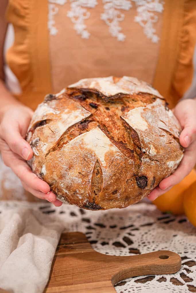 Woman holding a round loaf of sourdough cranberry orange bread, showing rustic golden crust and deep scoring detail.