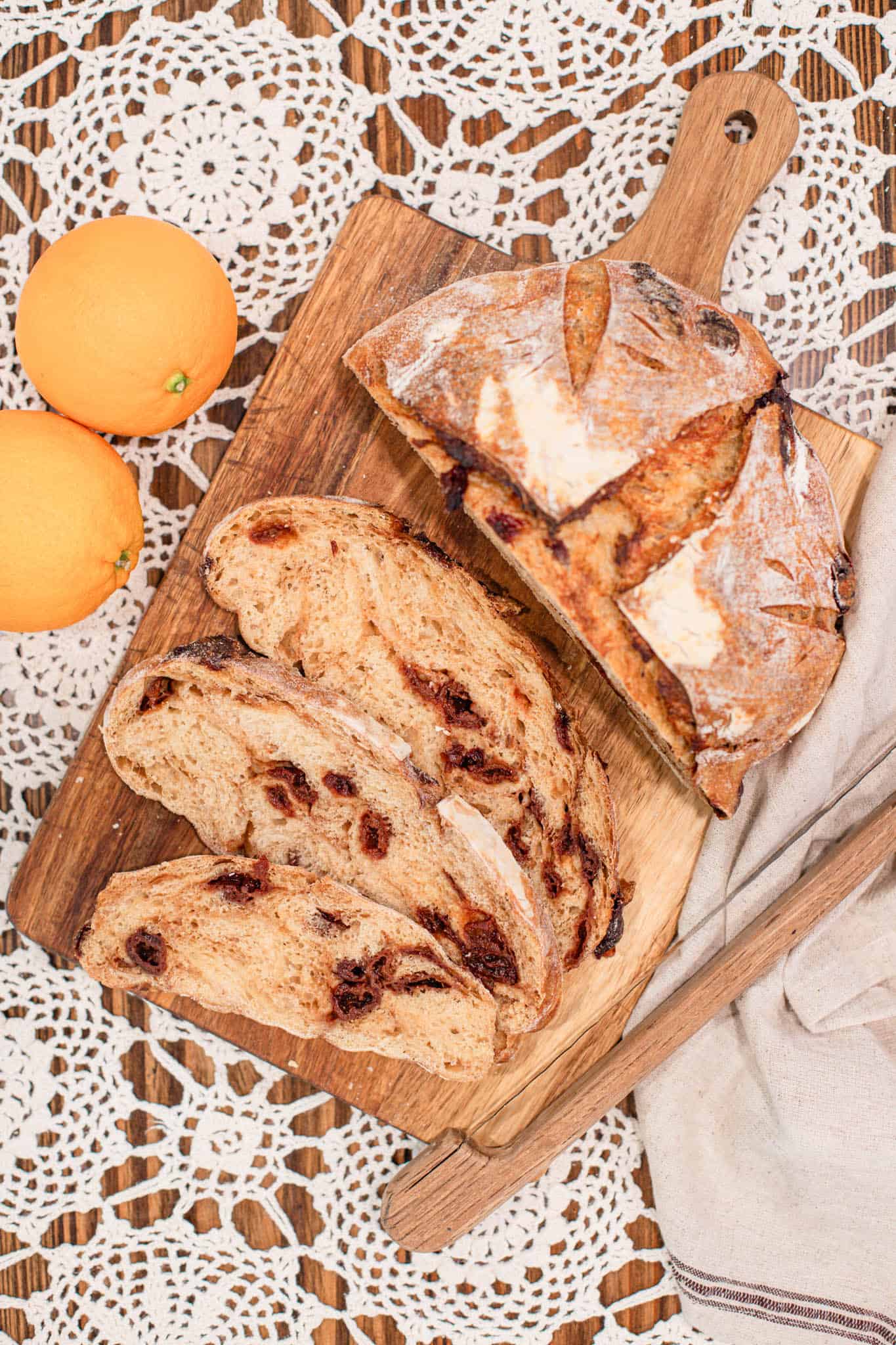 Overhead view of sliced sourdough cranberry orange bread on a wooden board beside fresh oranges and linen cloth.