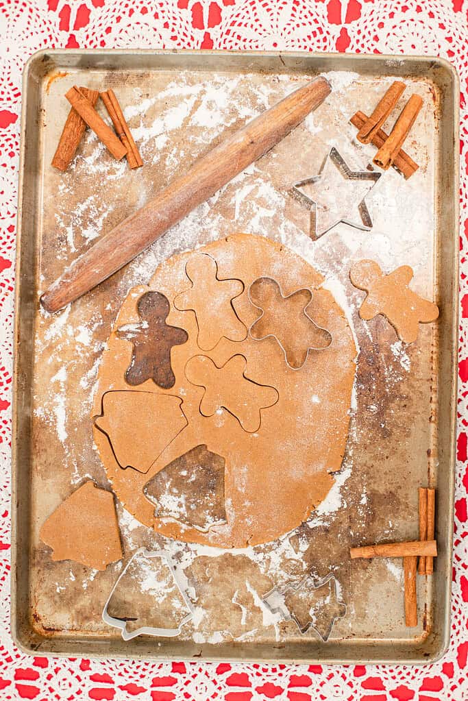 A sheet pan dusted with flour holds rolled gingerbread dough with several cookie shapes already cut out — gingerbread people, stars, bells, and trees. A wooden rolling pin rests across the top, and cinnamon sticks sit in little bundles around the edges. Everything is set on a red and white crocheted tablecloth that gives a cozy, handmade holiday feel.
