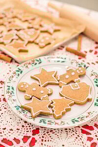 A holly-trimmed Christmas plate sits on the crocheted tablecloth and holds finished gingerbread cookies outlined with white icing. Shapes include stars, trees, gingerbread people, and a bell. Behind them is a blurred parchment-lined tray of more cookies, with a rolling pin and cinnamon sticks nearby for a warm holiday kitchen look.