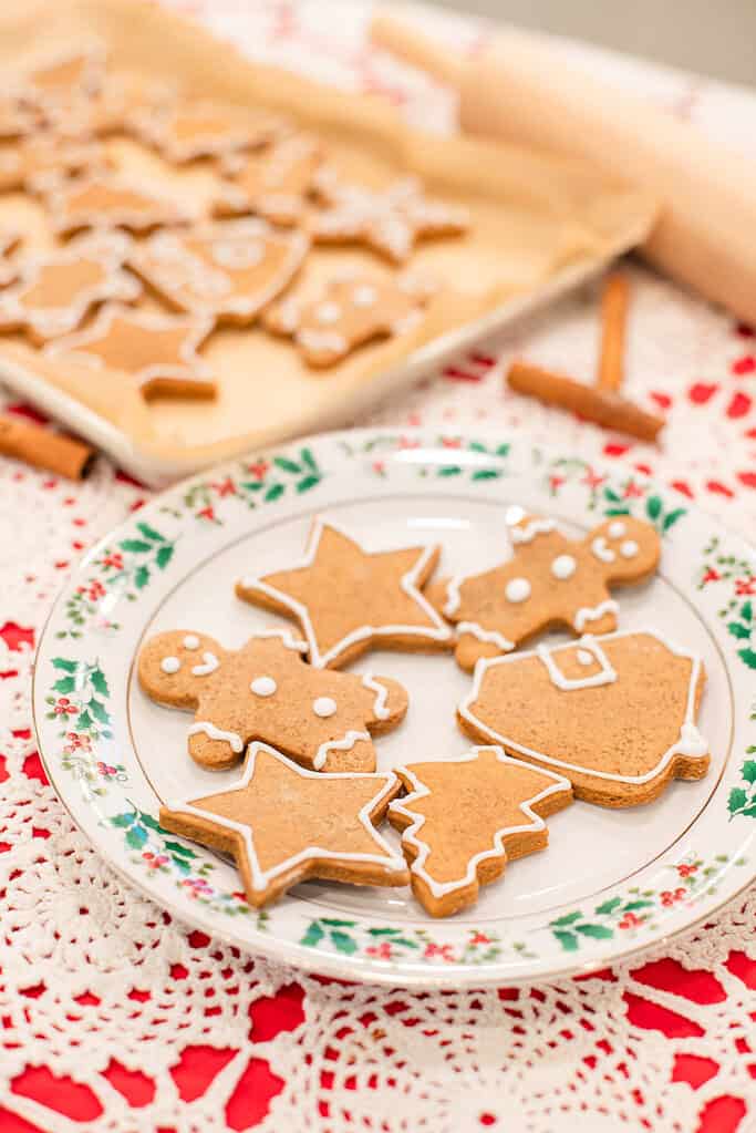 A holly-trimmed Christmas plate sits on the crocheted tablecloth and holds finished gingerbread cookies outlined with white icing. Shapes include stars, trees, gingerbread people, and a bell. Behind them is a blurred parchment-lined tray of more cookies, with a rolling pin and cinnamon sticks nearby for a warm holiday kitchen look.