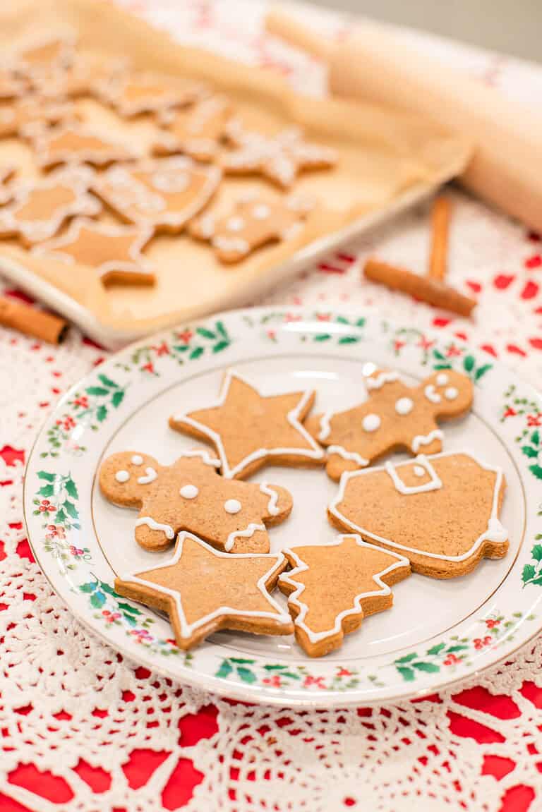 A holly-trimmed Christmas plate sits on the crocheted tablecloth and holds finished gingerbread cookies outlined with white icing. Shapes include stars, trees, gingerbread people, and a bell. Behind them is a blurred parchment-lined tray of more cookies, with a rolling pin and cinnamon sticks nearby for a warm holiday kitchen look.