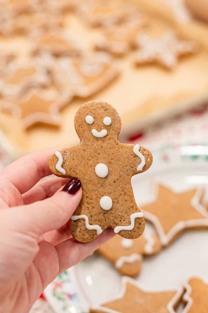 A hand with dark red nail polish holds a small gingerbread person outlined with white icing. The background is softly blurred with more cookies on a tray. The cookie’s texture, color, and details are clear — a friendly little smiling gingerbread figure.