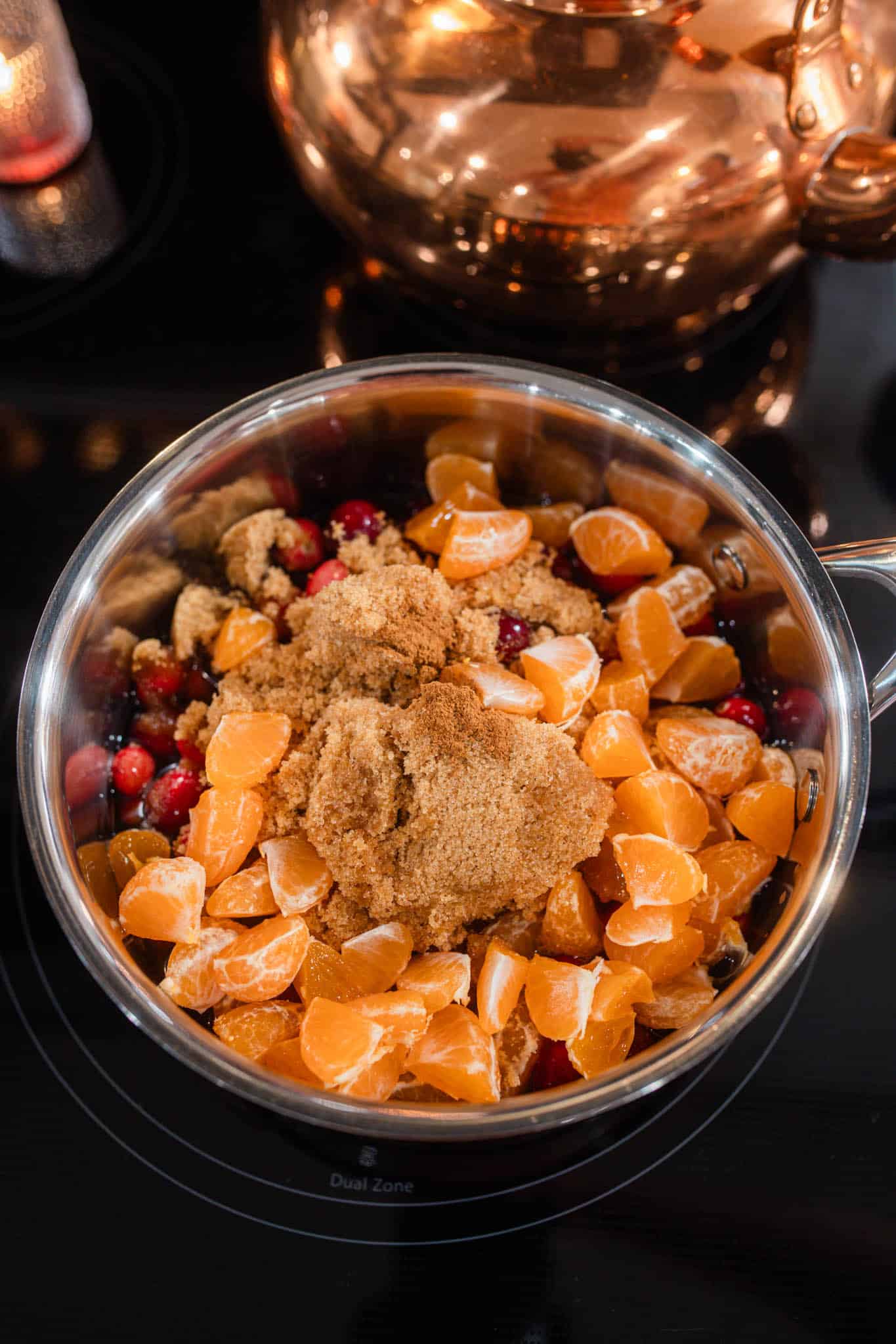 A saucepan filled with fresh cranberries, peeled orange segments, and a mound of brown sugar, ready to cook on the stovetop.