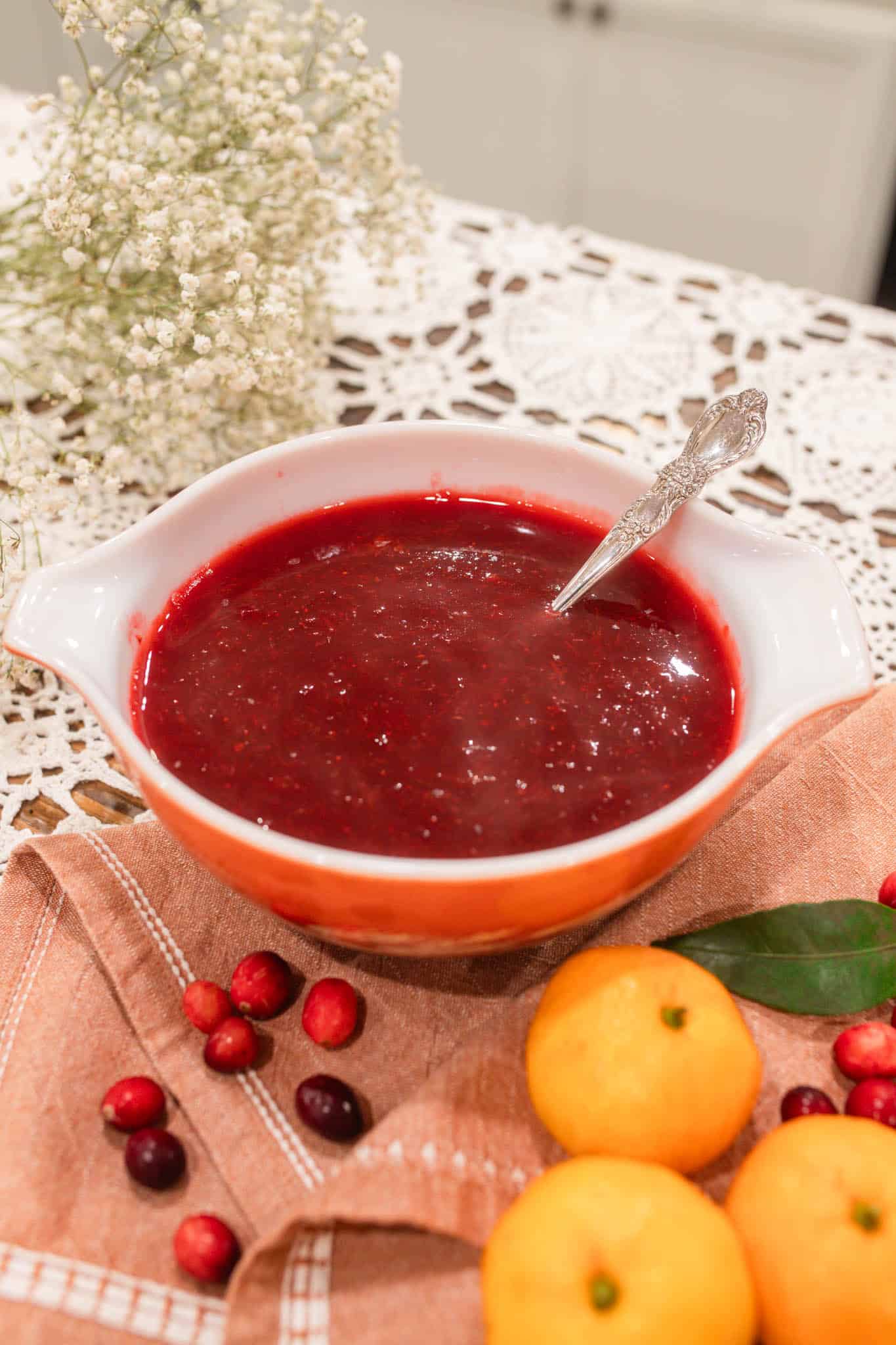 A serving bowl filled with cranberry orange sauce with a spoon inside, placed on a cloth with fresh cranberries and oranges nearby.