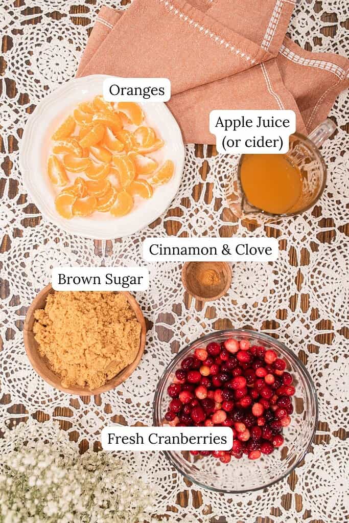 A top-down view of the ingredients for whole berry cranberry orange sauce arranged on a lace-covered table, including peeled orange segments, apple juice, brown sugar, fresh cranberries, and small bowls of cinnamon and clove.