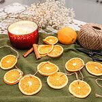Dried orange slice garland laid across a green textured cloth with a red candle, cinnamon sticks, whole oranges, and twine, styled as cozy handmade Christmas decor.