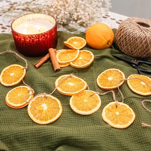 Dried orange slice garland laid across a green textured cloth with a red candle, cinnamon sticks, whole oranges, and twine, styled as cozy handmade Christmas decor.