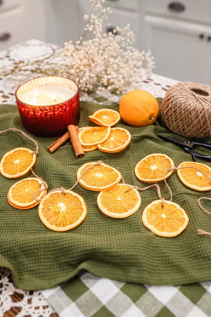 Dried orange slice garland laid across a green textured cloth with a red candle, cinnamon sticks, whole oranges, and twine, styled as cozy handmade Christmas decor.