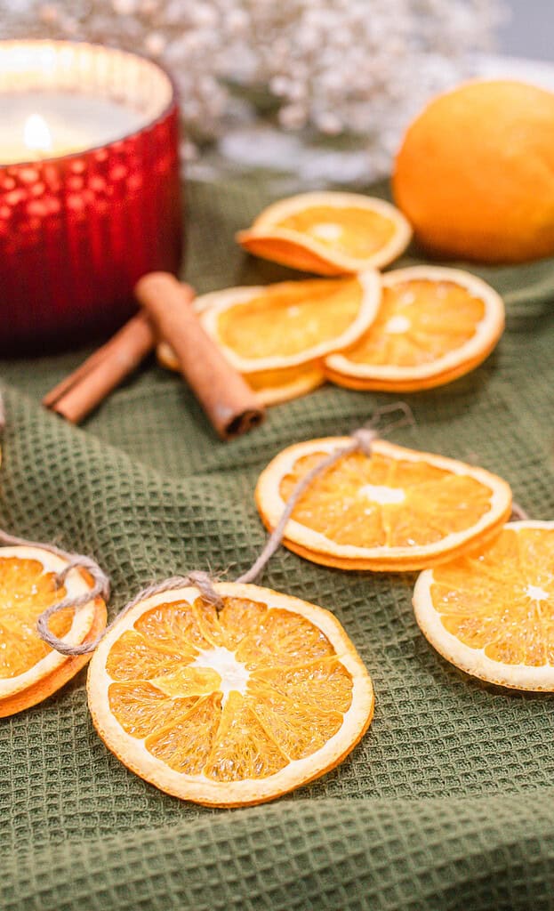 Dried orange slices tied with twine arranged on a green cloth alongside scissors, a ball of twine, cinnamon sticks, and a red candle, showing materials used to make an orange garland.