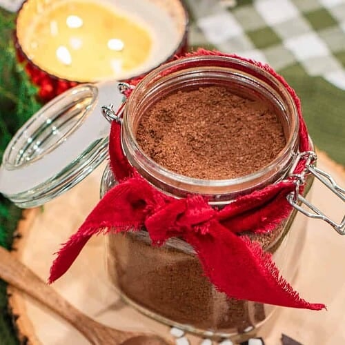 Homemade hot cocoa mix in a glass jar tied with a red ribbon, styled on a wood slice with a mug of hot chocolate topped with mini marshmallows.