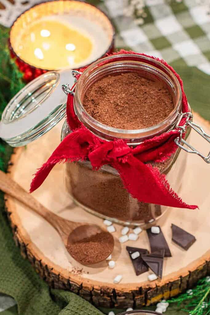 Homemade hot cocoa mix in a glass jar tied with a red ribbon, styled on a wood slice with a mug of hot chocolate topped with mini marshmallows.
