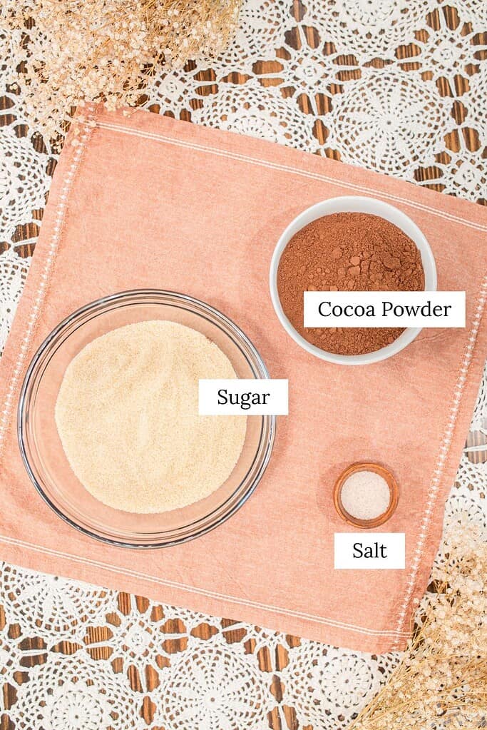 Hot cocoa mix ingredients laid out on a table, including granulated sugar, cocoa powder, and salt in separate bowls.