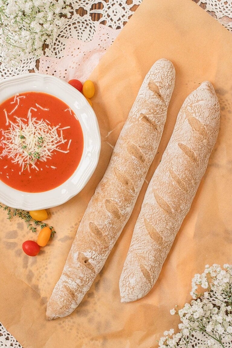 Two freshly baked sourdough baguettes made with freshly milled flour rest on parchment paper beside a white bowl of tomato soup topped with shredded cheese and herbs, styled with small tomatoes, greenery, and delicate white flowers on a rustic lace tablecloth.