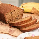Close-up of a sliced freshly milled flour banana bread loaf on butcher paper with a banana in the background, showing a moist golden-brown crumb with visible walnut pieces, one slice resting on a vintage floral china plate.