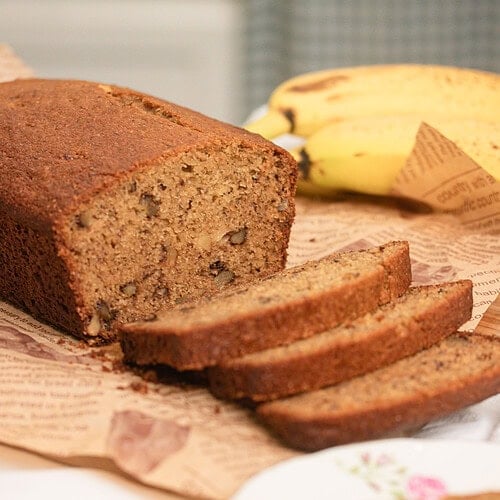 Close-up of a sliced freshly milled flour banana bread loaf on butcher paper with a banana in the background, showing a moist golden-brown crumb with visible walnut pieces, one slice resting on a vintage floral china plate.
