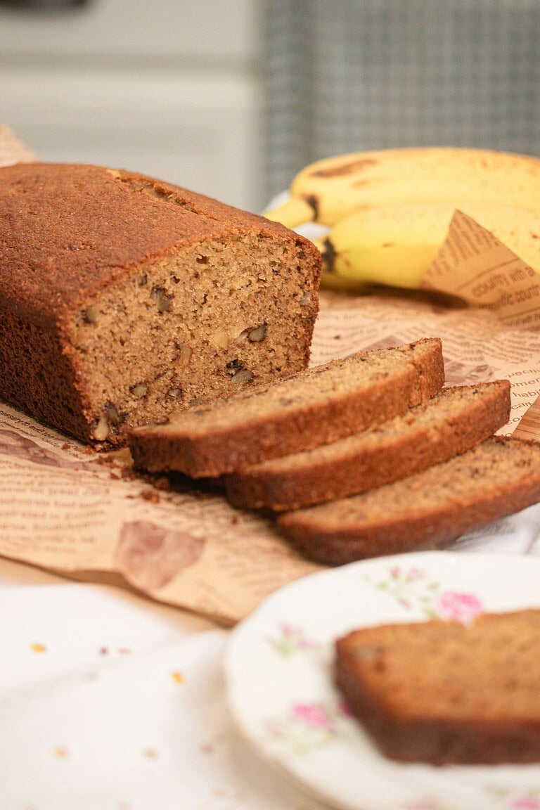 Close-up of a sliced freshly milled flour banana bread loaf on butcher paper with a banana in the background, showing a moist golden-brown crumb with visible walnut pieces, one slice resting on a vintage floral china plate.