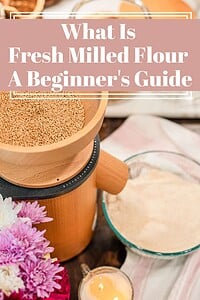 A wooden grain mill loaded with whole wheat berries next to a glass bowl of freshly milled flour, styled with pink and white chrysanthemums and a striped linen, with a title text overlay reading What Is Fresh Milled Flour A Beginner's Guide.
