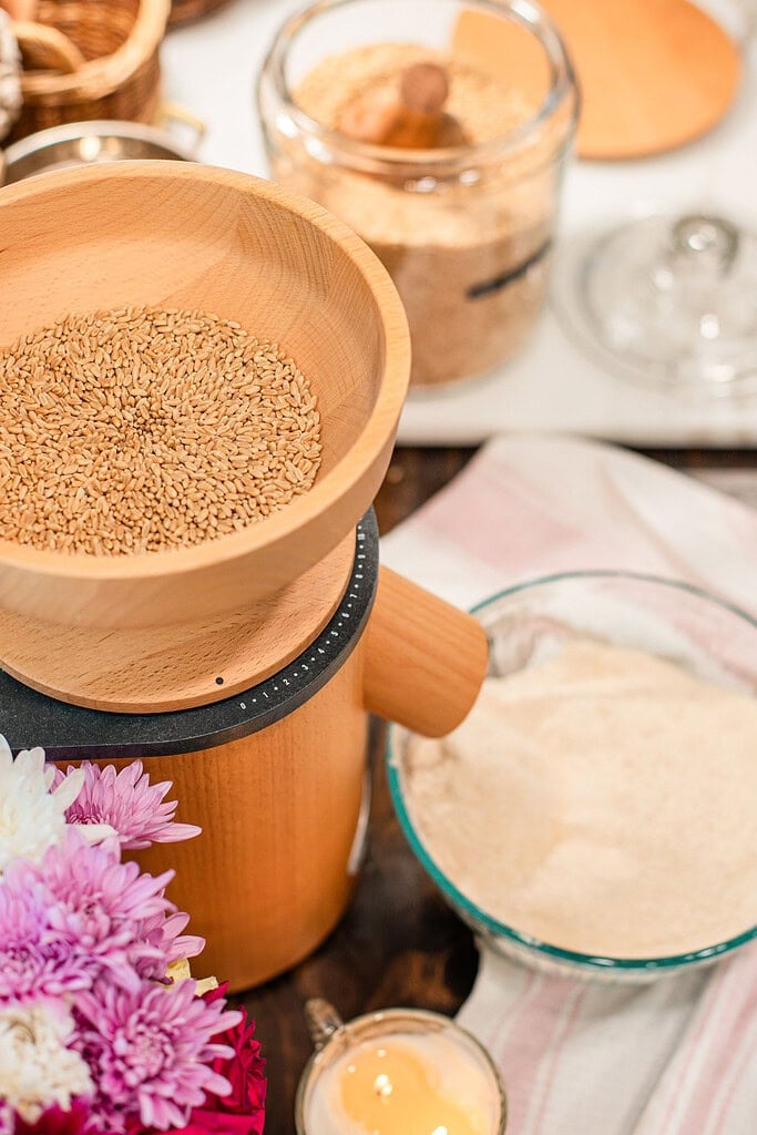 A wooden grain mill with whole wheat berries in the hopper next to a glass bowl of freshly milled flour, styled with a glass jar of flour, pink and white chrysanthemums, and a striped linen in a cottage kitchen.