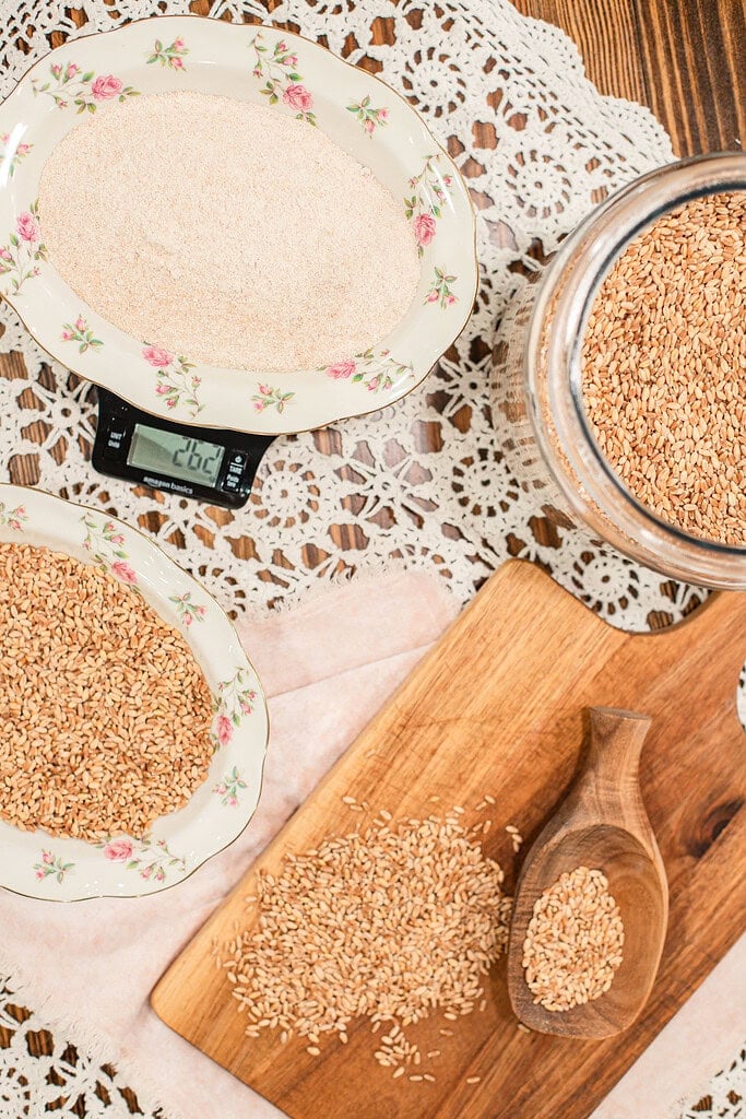 Overhead view of whole wheat berries and freshly milled flour on vintage rose-rimmed china plates next to a kitchen scale, with a glass jar of wheat berries and a wooden scoop on a cutting board on a lace doily.