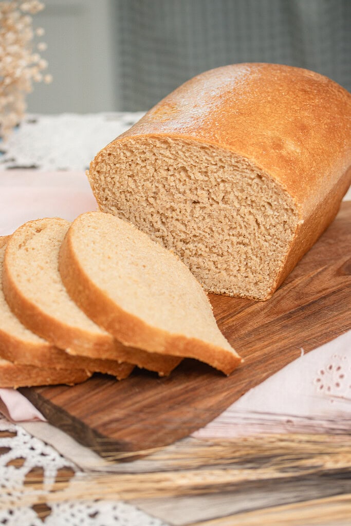 A partially sliced loaf of fresh milled flour sandwich bread on a wood cutting board showing the soft golden crumb, styled on a dusty rose linen with a lace doily and dried wheat stems in a cottage kitchen.