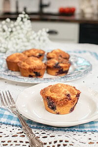 A single golden bakery-style blueberry muffin made with freshly milled flour on a small white plate with a silver fork, with more muffins on a blue transferware platter and white baby's breath flowers in the background inside a cottage kitchen.
