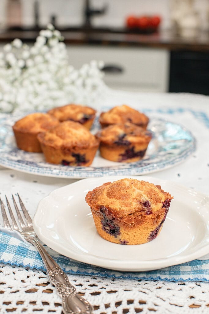 A single golden bakery-style blueberry muffin made with freshly milled flour on a small white plate with a silver fork, with more muffins on a blue transferware platter and white baby's breath flowers in the background inside a cottage kitchen.
