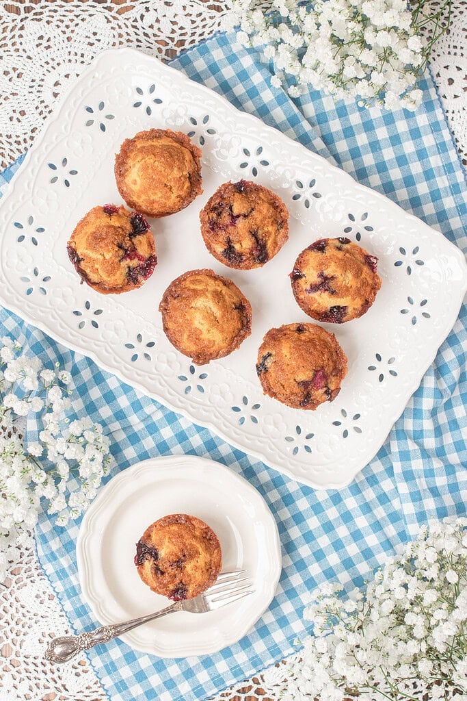 Overhead view of six bakery-style blueberry streusel muffins made with freshly milled flour arranged on a white pierced ceramic tray, with one muffin on a small white plate with a fork, surrounded by white baby's breath flowers on a blue gingham cloth and white lace doily.