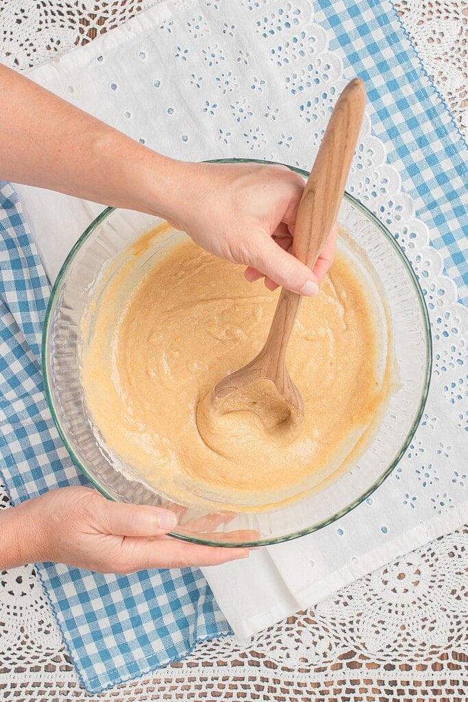 Overhead view of hands stirring thick golden muffin batter in a large glass bowl with a wooden spoon, set on a blue gingham cloth and white eyelet linen.