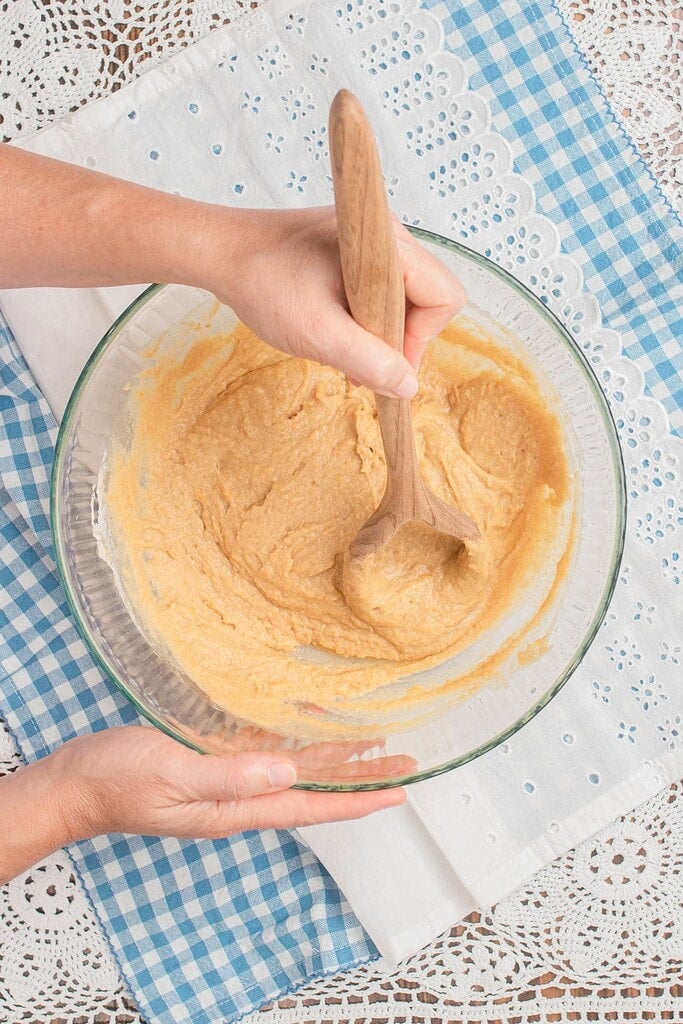 Overhead view of hands folding thick freshly milled flour muffin batter in a large glass bowl with a wooden spoon, set on a blue gingham cloth and white eyelet linen.