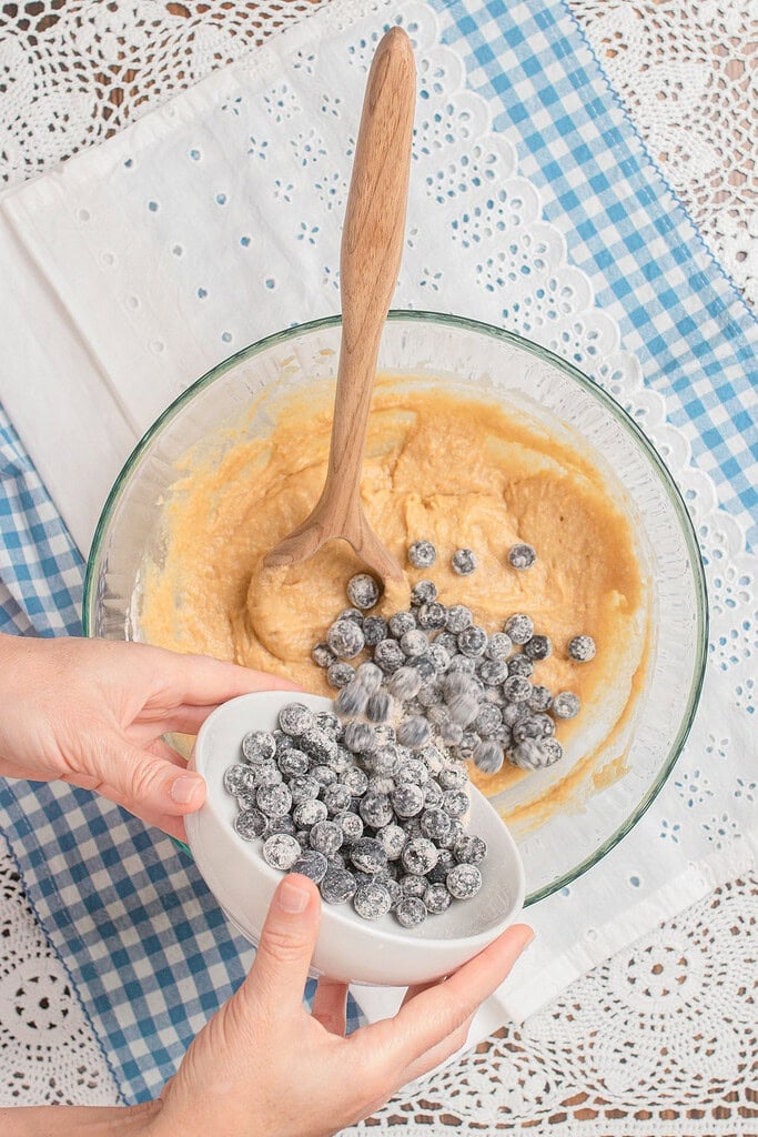 Overhead view of hands pouring flour-dusted blueberries from a small white bowl into thick golden muffin batter in a large glass bowl, set on a blue gingham cloth and white lace doily.