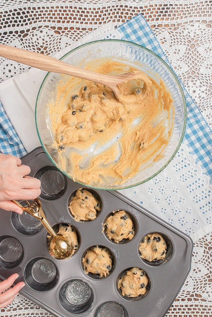 Overhead view of hands using a gold cookie scoop to portion freshly milled flour blueberry muffin batter into a gray nonstick Wilton muffin tin, with the glass mixing bowl of remaining batter nearby on a white lace doily.